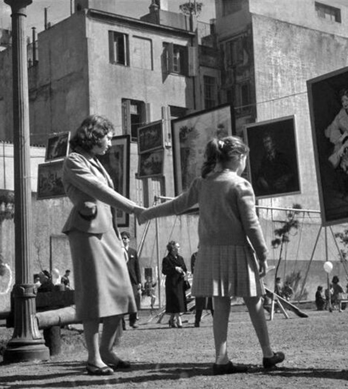 Una mujer y una niña de la mano frente a cuadros colgados al aire libre en una plaza porteña.