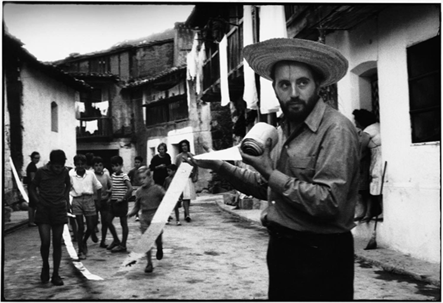 Greco con un rollo de papel en una calle de pueblo, rodeado de vecinos.