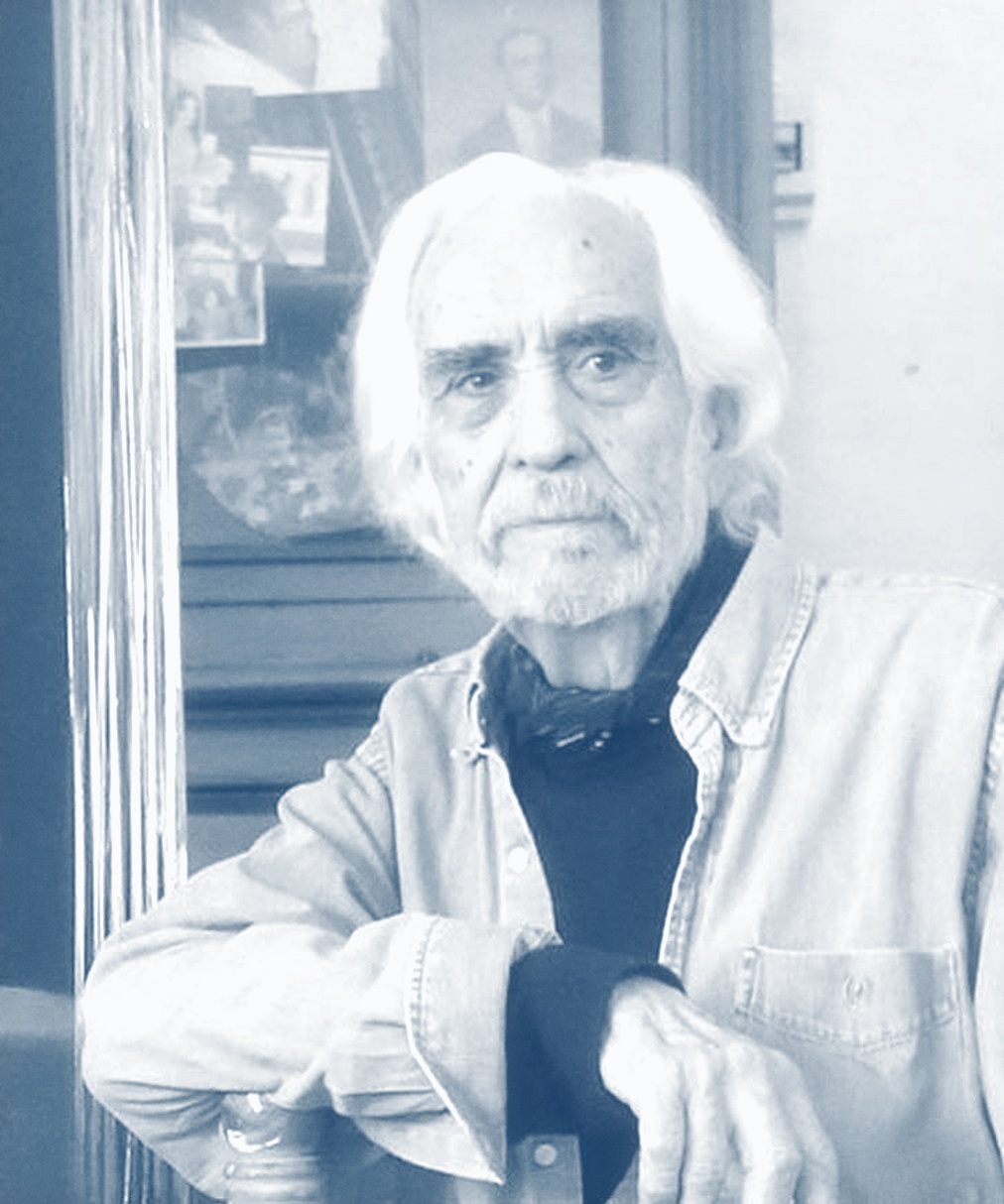 Carlos Alonso, de pelo y barba blancos y camisa de jean, sentado junto a una de sus pinturas.
