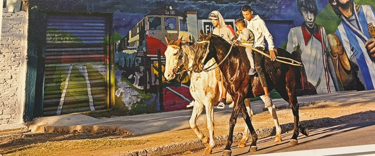 «Conurbano Style», de Marcos López junto a The Walking Conurban: tres chicos a caballo pasan frente a un mural con un tren y dos retratos de Maradona.