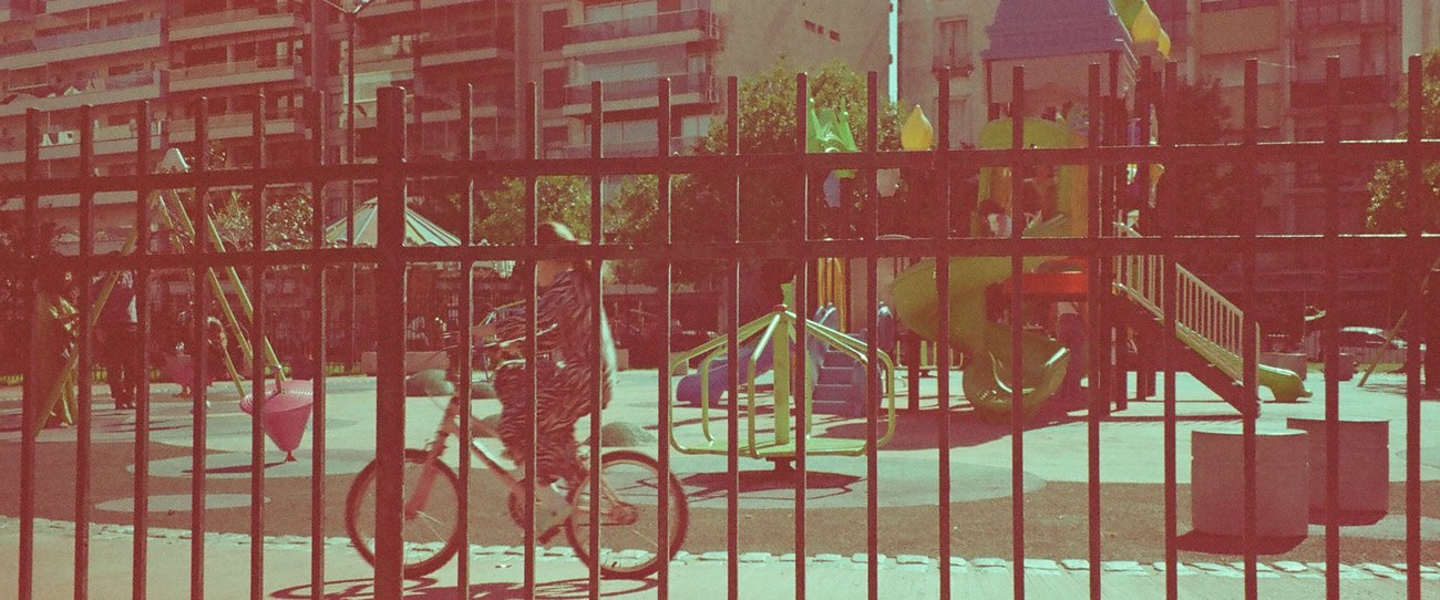 Juegos de plaza detrás de una reja, con torres de departamentos al fondo y una persona en bicicleta pasando.